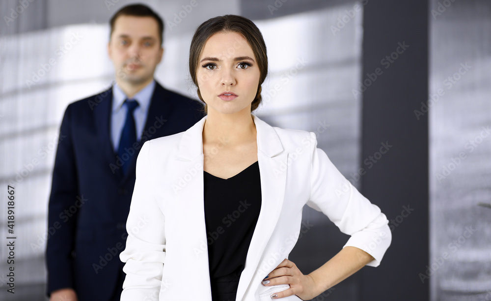 Young beautiful businesswoman is standing in an office with a male colleague at the background. Concept of success in a business Young beautiful businesswoman is standing in an office with a male colleague at the background. Concept of success in a business