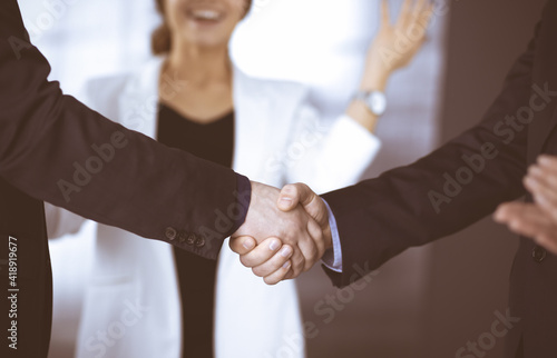 Businessmen are shaking hands while standing with colleagues applauding in office, close-up. Business success concept