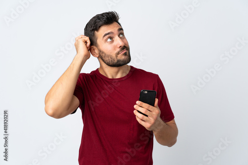 Young handsome man with beard using mobile phone isolated on white background having doubts and with confuse face expression © luismolinero