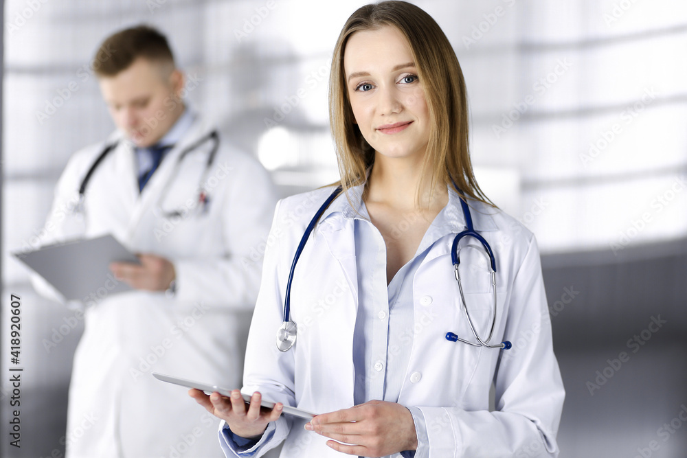 Professional woman doctor with a stethoscope is using a computer tablet, while she is standing together with her colleague in a clinic. Young doctors at work in a hospital. Medicine and healthcare Professional woman doctor with a stethoscope is using a computer tablet, while she is standing together with her colleague in a clinic. Young doctors at work in a hospital. Medicine and healthcare