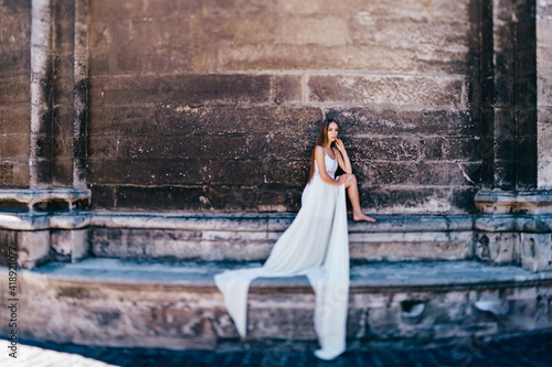 Young romantic elegant girl in long white flowy dress posing over stone ancient wall © benevolente