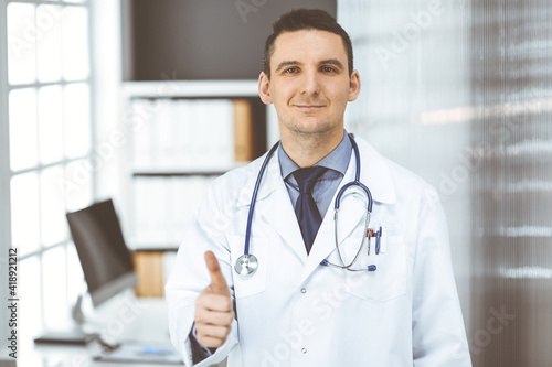 Male doctor standing with thumbs up sign in clinic near his working place. Perfect medical service in hospital. Medicine and healthcare concept