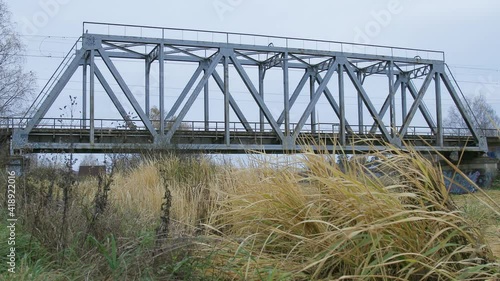 Suburban train is crossing the small bridge over water from right to left in countryside-like location in Russia. Autumn cloudy weather. Side view