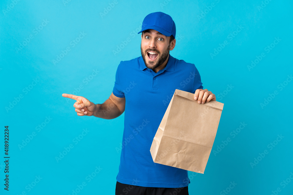 Delivery man with beard isolated on blue background surprised and pointing finger to the side Delivery man with beard isolated on blue background surprised and pointing finger to the side