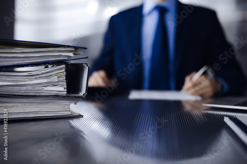 Binders with papers are waiting to be processed by man accountant in blue blazer staying at home during covid pandemic. Taxes and audit concept. Internal Revenue Service inspector checking financial