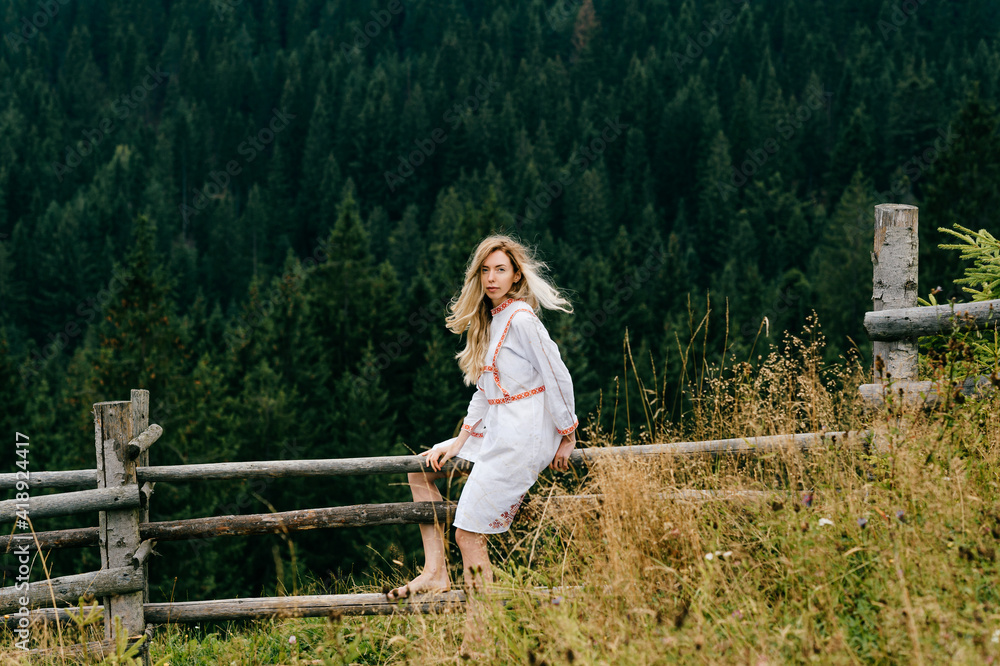 Young attractive blonde girl in white dress with embroidery sitting on wooden fence over picturesque forest landscape Young attractive blonde girl in white dress with embroidery sitting on wooden fence over picturesque forest landscape