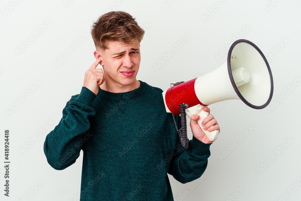 Young caucasian man holding a megaphone isolated on white background covering ears with hands. Young caucasian man holding a megaphone isolated on white background covering ears with hands.