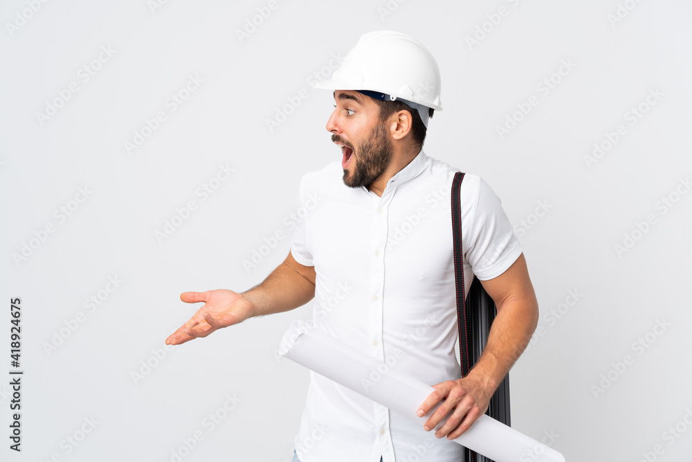 Young architect man with helmet and holding blueprints isolated on white background with surprise expression while looking side