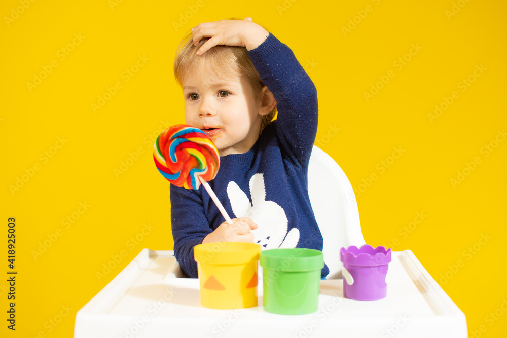 Cute little boy holding colorful lollipop while against sitting on chair over yellow background. Cute little boy holding colorful lollipop while against sitting on chair over yellow background.