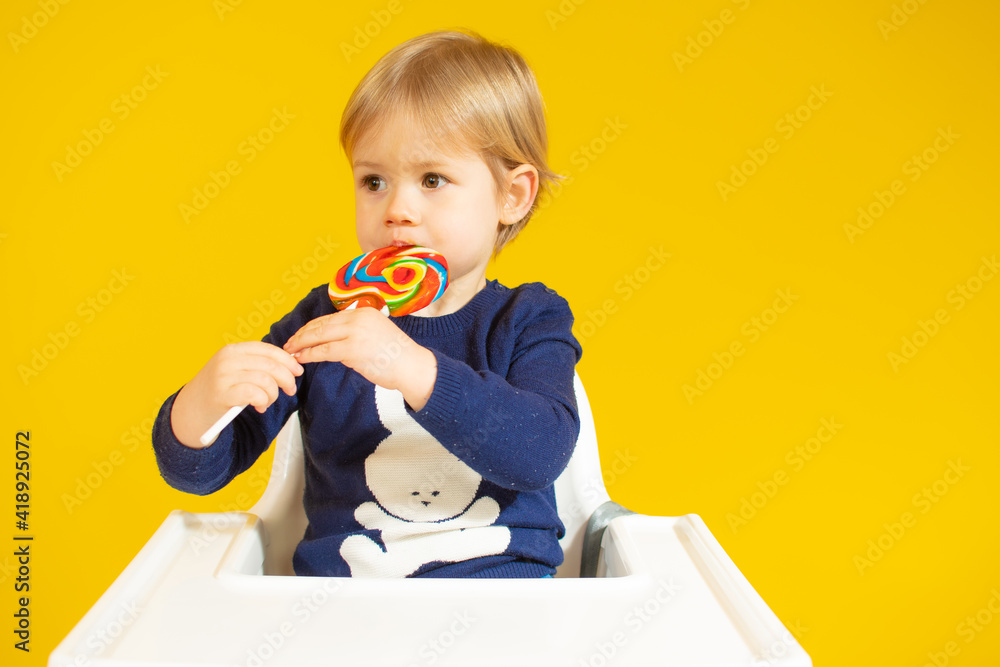 Cute little boy eating colorful lollipop while against sitting on chair over yellow background. Cute little boy eating colorful lollipop while against sitting on chair over yellow background.