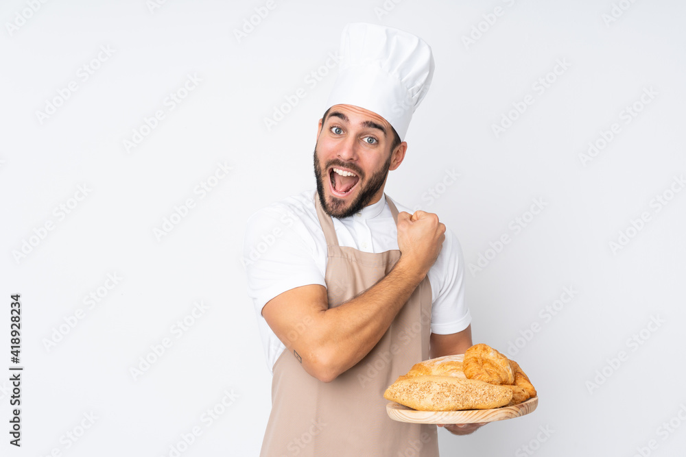 Male baker holding a table with several breads isolated on white background celebrating a victory Male baker holding a table with several breads isolated on white background celebrating a victory