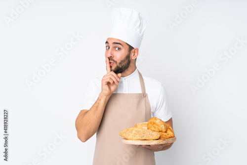 Male baker holding a table with several breads isolated on white background doing silence gesture