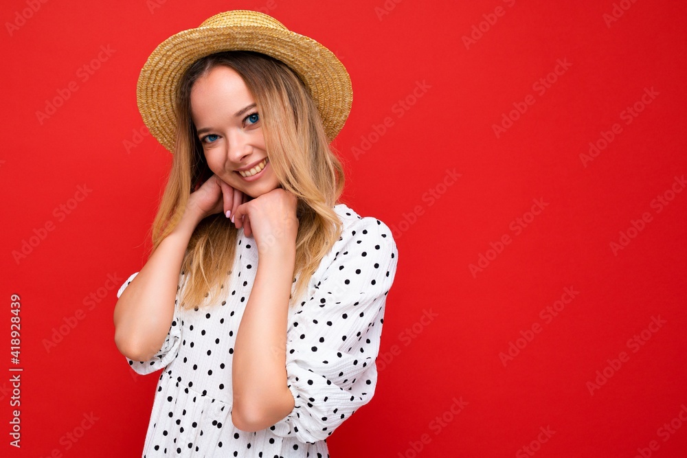 Portrait of young beautiful smiling hipster blonde woman in trendy summer dress and straw hat. Sexy carefree female person posing isolated near red wall in studio. Positive model with natural makeup Portrait of young beautiful smiling hipster blonde woman in trendy summer dress and straw hat. Sexy carefree female person posing isolated near red wall in studio. Positive model with natural makeup