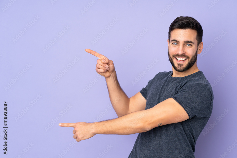 Caucasian handsome man pointing finger to the side and presenting a product over isolated purple background Caucasian handsome man pointing finger to the side and presenting a product over isolated purple background