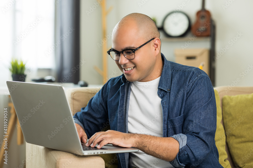 Indian man using laptop on sofa in living room texting on laptop sending message or chatting with online social media. Indian man using laptop on sofa in living room texting on laptop sending message or chatting with online social media.