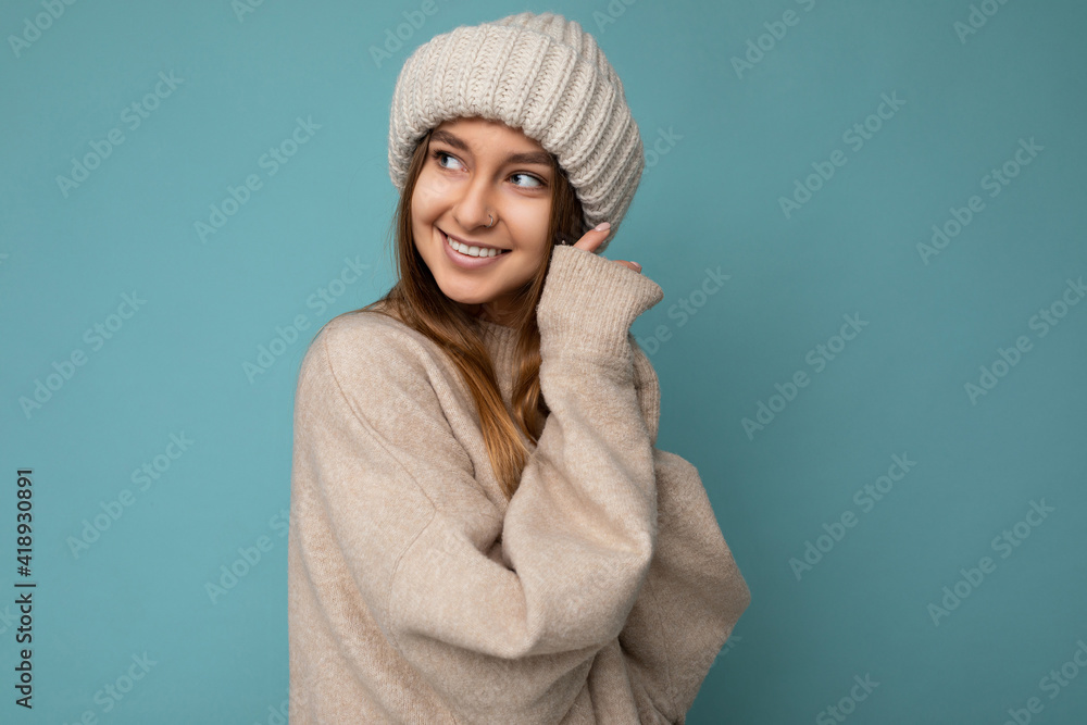 Shot of attractive smiling amazing happy young dark blonde woman standing isolated on blue background wall wearing beige warm sweater and winter beige hat looking to the side