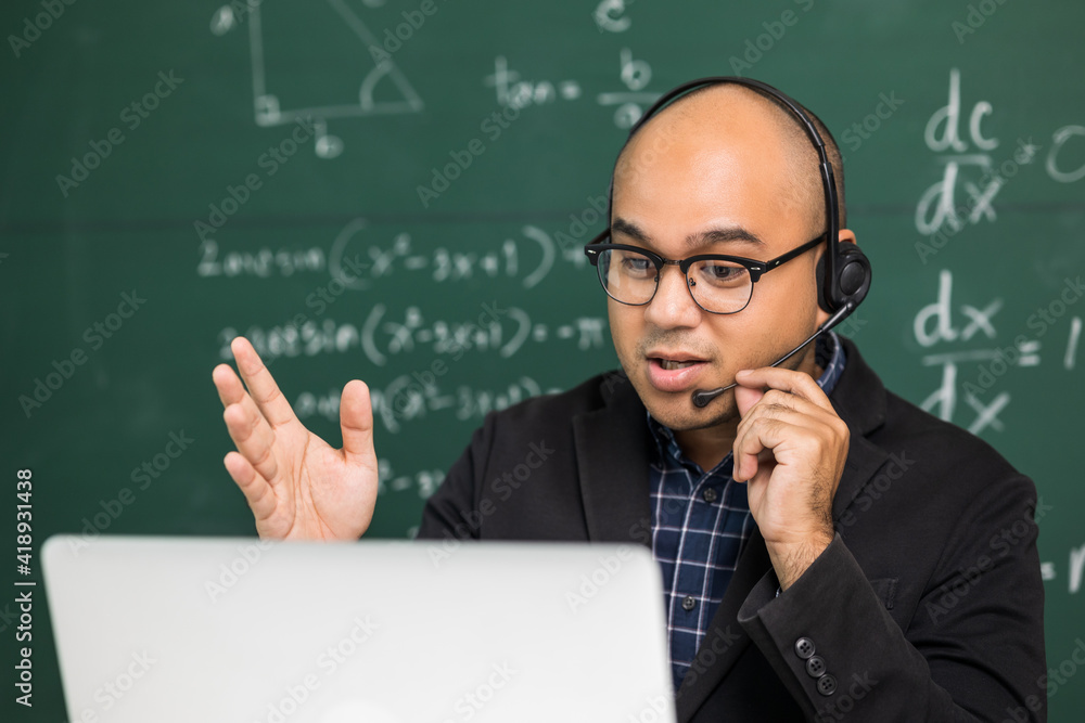 Indian young teacher man sitting wearing headset teaching online video conference live stream by laptop. Asian teacher teaching mathematics class webinar online for students learning.