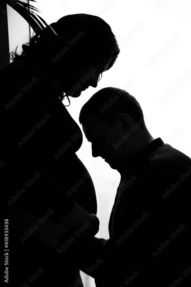 silhouette of future parents. father kneels and looks at the belly of his pregnant wife. black and white photograph of a pregnant woman. future parents.