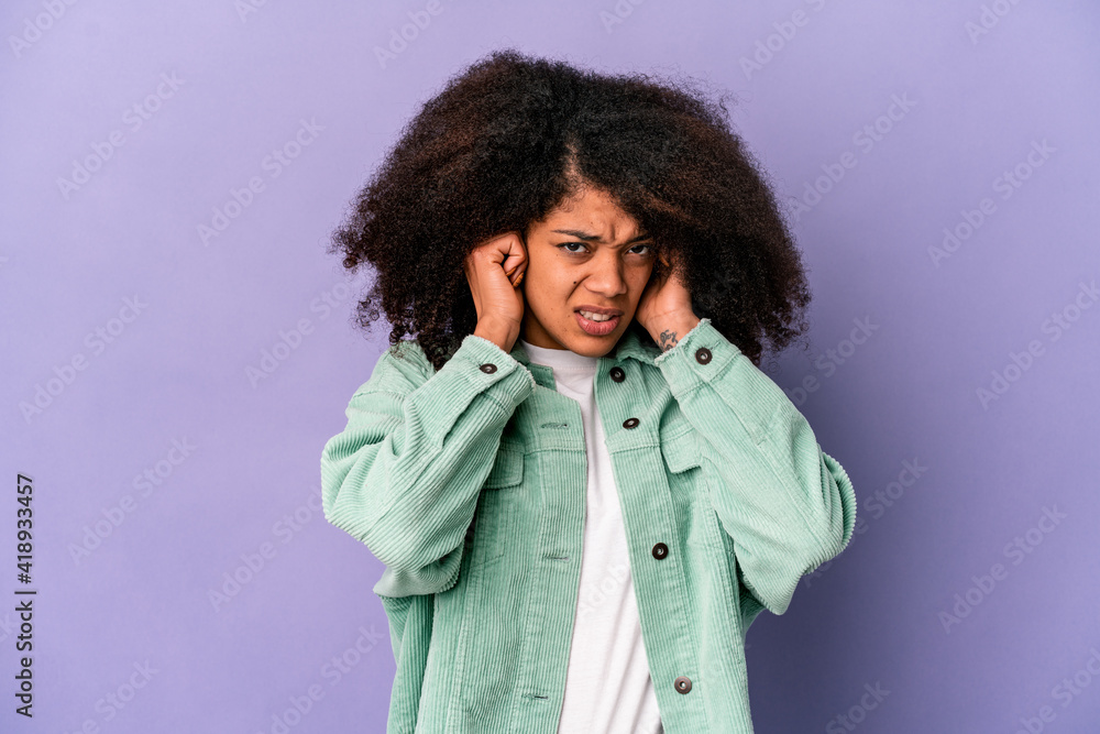 Young african american curly woman isolated on purple background covering ears with fingers, stressed and desperate by a loudly ambient. Young african american curly woman isolated on purple background covering ears with fingers, stressed and desperate by a loudly ambient.