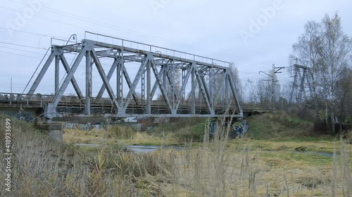Suburban train is crossing the small bridge over water  in countryside-like location in Russia. Autumn cloudy weather. 