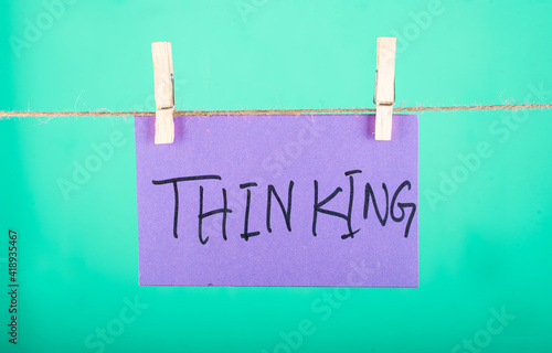 Thinking word written on a Purple color sticky note hanging with a wire in a Cyan background.