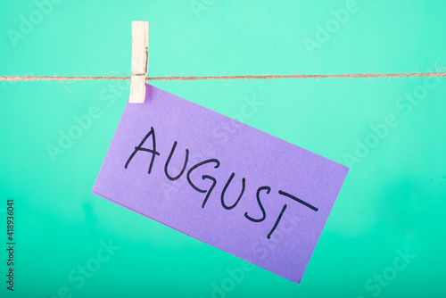 August word written on a Purple color sticky note hanging with a wire in a Cyan background.
