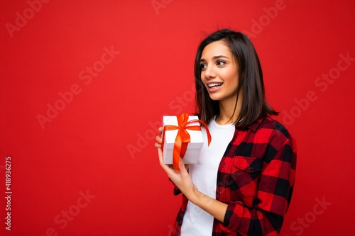 Beautiful happy young brunette woman isolated over colourful background wall wearing stylish casual clothes holding gift box and looking to the side © Ivan Traimak