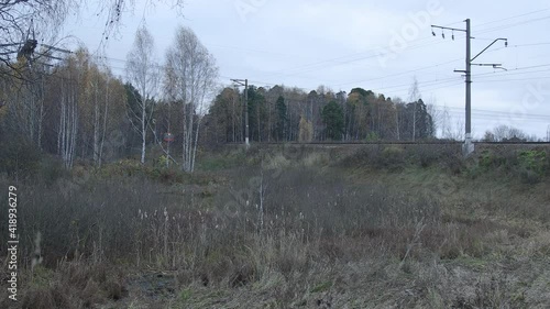 Suburban train is appearing from the woods and crossing the small bridge over water in countryside-like location in Russia. Autumn cloudy weather. Tracking shot from left to right