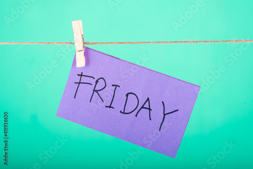 Friday word written on a Purple color sticky note hanging with a wire in a Cyan background.