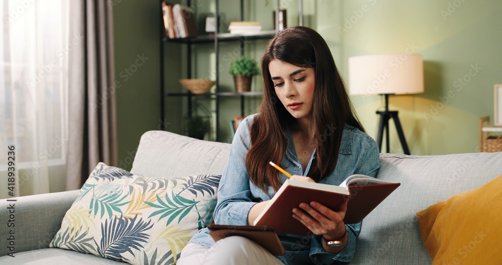 Naklejka premium Beautiful caucasian girl sitting at home on couch and writing in notebook lecture concept looking at tablet.