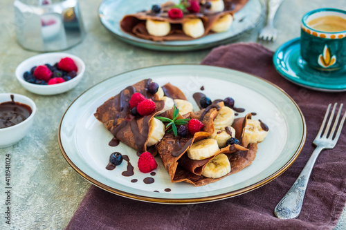 Thin chocolate pancakes stuffed with bananas, raspberries and blueberries, poured with chocolate sauce, on a gray ceramic plate on a light concrete background. Pancake recipes.