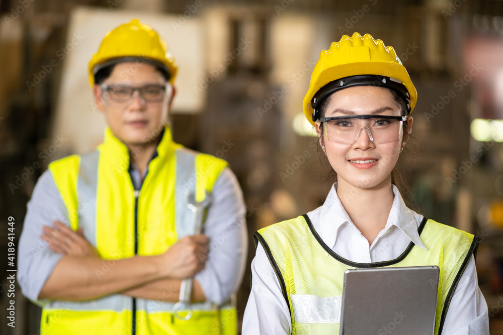 Portrait of confident Asian beautiful woman engineering crossing arm and holding tablet with blurry engineer man on behind in the factory. Portrait of confident Asian beautiful woman engineering crossing arm and holding tablet with blurry engineer man on behind in the factory.