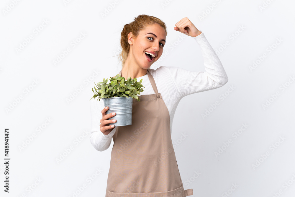 Young blonde gardener woman girl holding a plant over isolated white background making strong gesture Young blonde gardener woman girl holding a plant over isolated white background making strong gesture