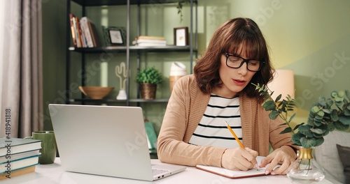 Portrait of happy smiling positive Caucasian young woman in glasses browsing online searching internet tapping on laptop and writing down information in notebook planner while learning sitting at desk © VAKSMANV