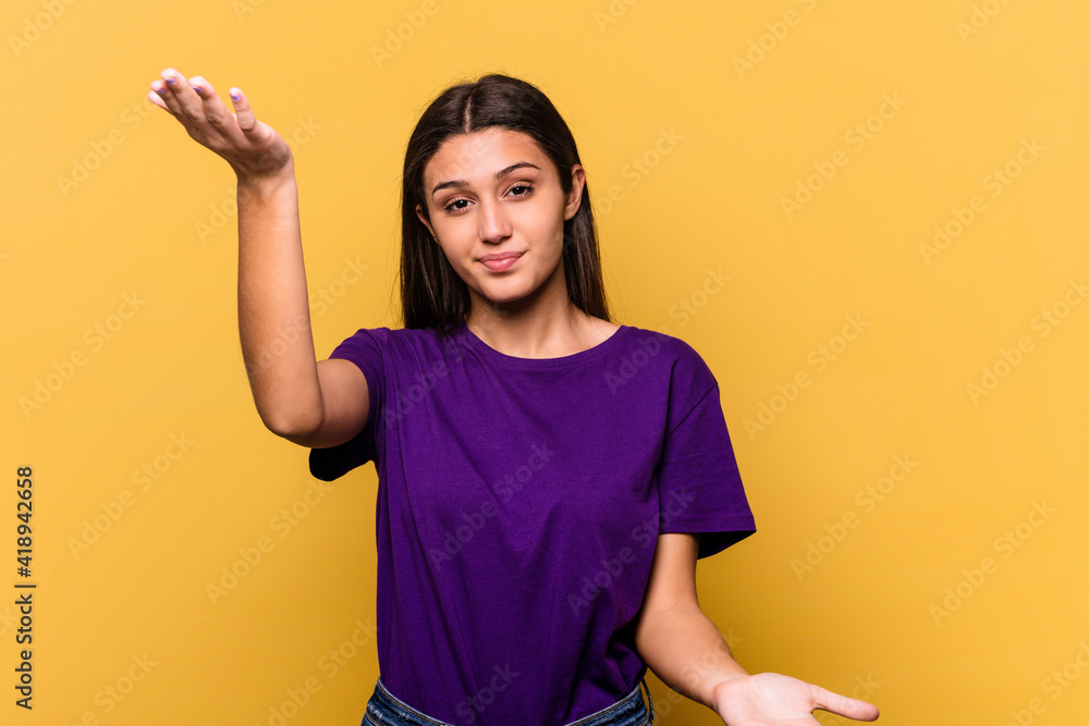 Young Indian woman isolated on yellow background makes scale with arms, feels happy and confident. Young Indian woman isolated on yellow background makes scale with arms, feels happy and confident.
