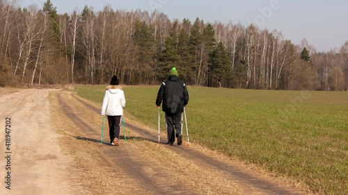  Suburbs of Grodno. Belarus. Spring landscape..Country road and two - a man and a woman engaged in Nordic walking, field, forest, blue spring sky with white clouds. © Volha