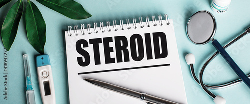 In a white notebook on a blue background, near a sheet of shefflers, a stethoscope, a syringe and an electronic thermometer, the word STEROID is written. Medical concept photo