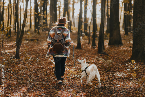 Stylish woman with backpack hiking with white dog in sunny autumn woods. Cute swiss shepherd puppy