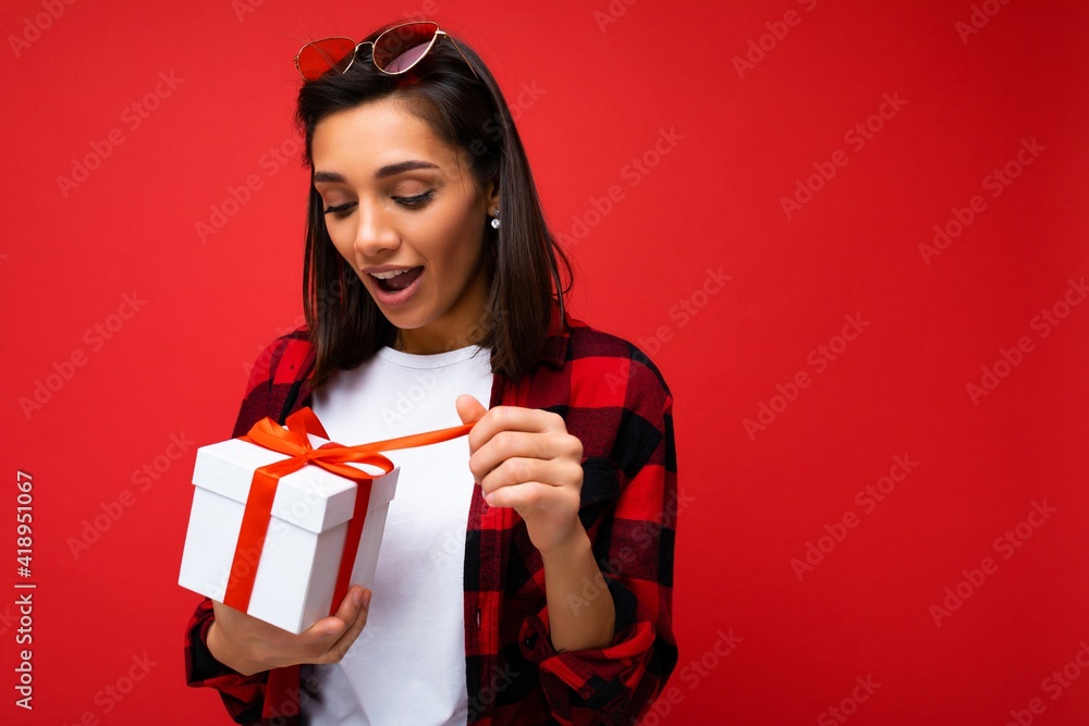 Charming positive surpeised young brunet woman isolated on red background wall wearing white casual t-shirt and stylish red and black shirt holding white gift box with red ribbon and unboxing surprise Charming positive surpeised young brunet woman isolated on red background wall wearing white casual t-shirt and stylish red and black shirt holding white gift box with red ribbon and unboxing surprise