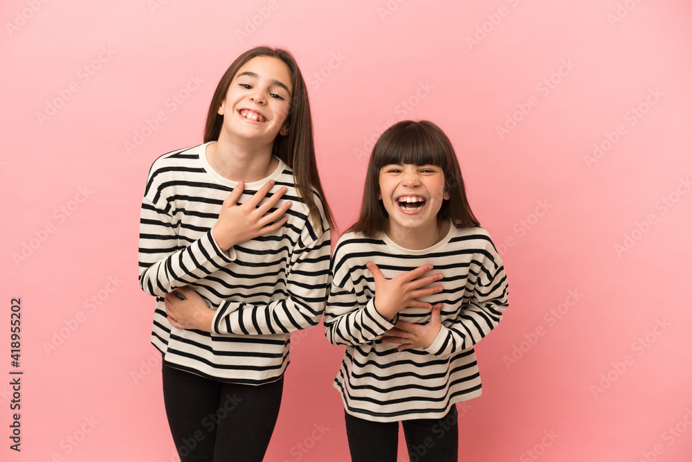 Little sisters girls isolated on pink background smiling a lot while putting hands on chest Little sisters girls isolated on pink background smiling a lot while putting hands on chest