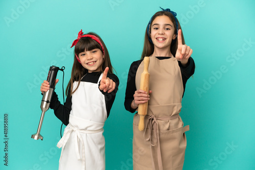 Little sisters cooking at home isolated on blue background showing and lifting a finger
