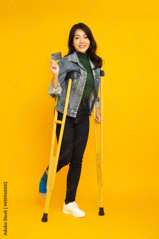 Portrait of a happy young Asian woman using a crutch and showing credit card on hand isolated on yellow background, Foot injury and Personal accident concept