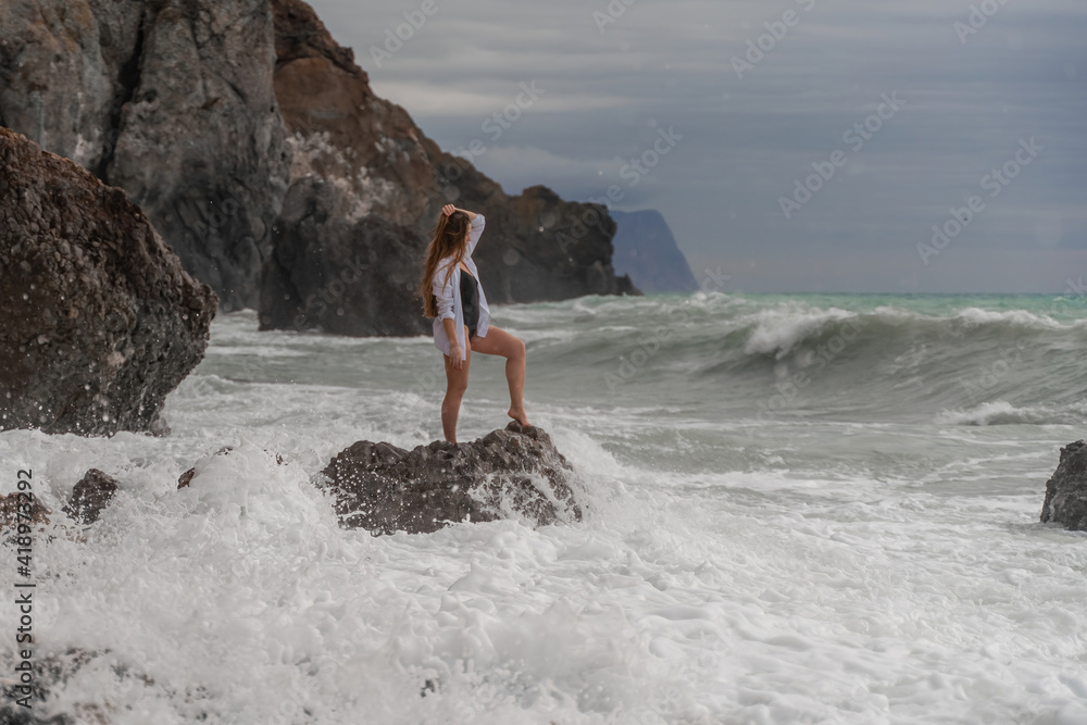 A beautiful girl in a white shirt and black swimsuit stands on a rock, big waves with white foam. A cloudy stormy day at sea, with clouds and big waves hitting the rocks.