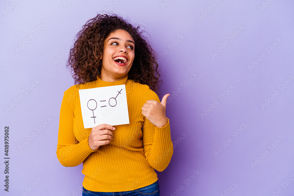 Young African American woman holding a gender equality placard isolated on purple background points with thumb finger away, laughing and carefree. Young African American woman holding a gender equality placard isolated on purple background points with thumb finger away, laughing and carefree.