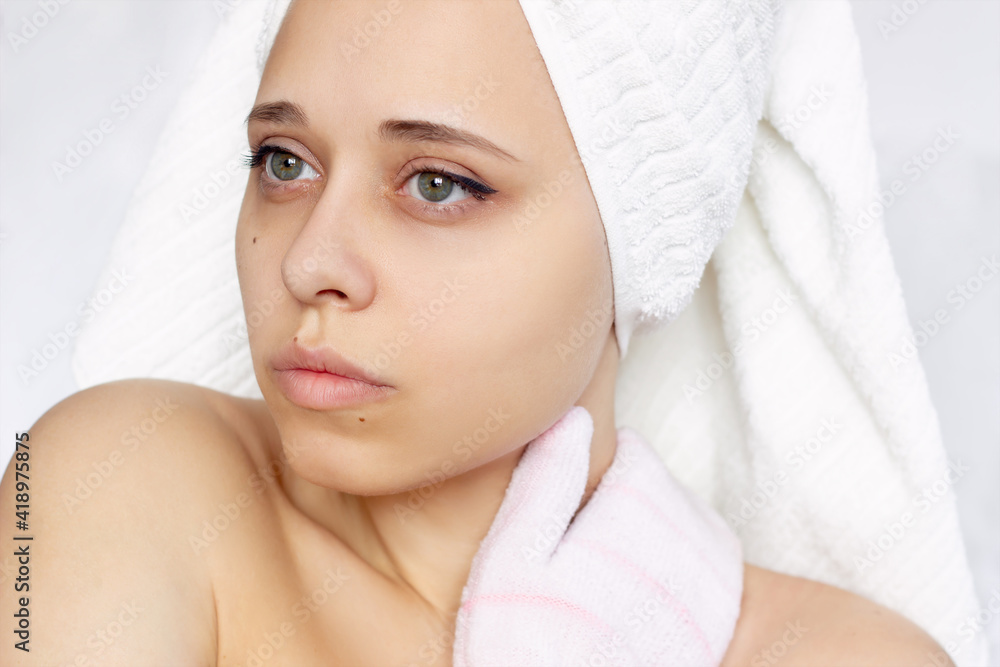 A young beautiful caucasian woman with a white towel on her head after a shower massaging her neck with an exfoliating hydro glove. Skin care, cosmetology A young beautiful caucasian woman with a white towel on her head after a shower massaging her neck with an exfoliating hydro glove. Skin care, cosmetology