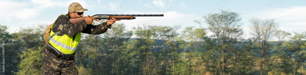 Fototapeta premium A hunter with a gun in his hands in hunting clothes in the autumn forest in search of a trophy. A man stands with weapons and hunting dogs tracking down the game.