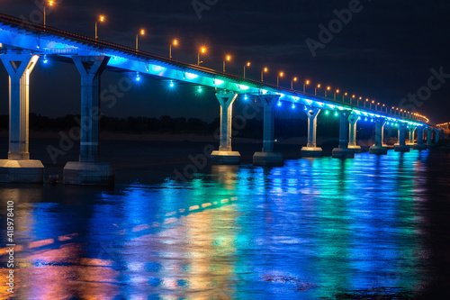 Side view of colourful bridge illuminated with blue and green color lights at the night. Bridge stands on Volga river in Russia. Colourful light is reflected in the water.
