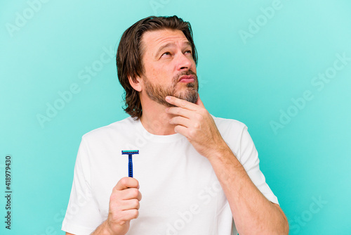 Middle age dutch man holding a razor blade isolated on blue background looking sideways with doubtful and skeptical expression.