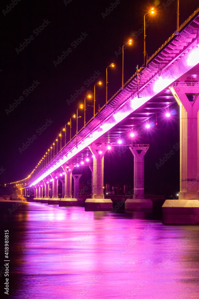 Side view of colourful bridge illuminated with pink color lights at the night. Bridge stands on Volga river in Russia. Pink light is reflected in the water.