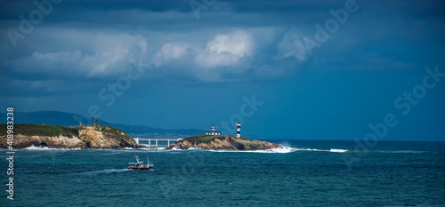 Un faro en una pequeña isla unida a tierra mediante un puente, un barco pesquero de color negro, nubes de tormenta y el mar cantábrico en la costa española